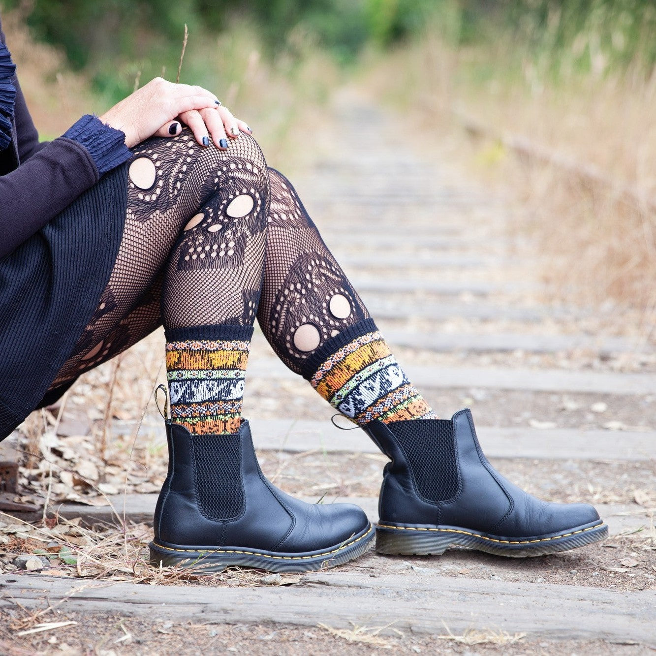 Side angle of a woman resting on a railroad tie, hands on her knees, skull tights tucked into black boots cuffed with Spooky Season Sweater socks in Black and Orange featuring blue, mustard, and cream knit patterning. [lf]