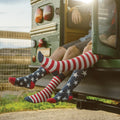 Two friends dangle their legs from the open back of a vintage green school bus at golden hour, their knee-high American Flag XL socks in VINTAGE BLUE showing red and white stripes meeting star-spangled navy panels above red toes. [lf]