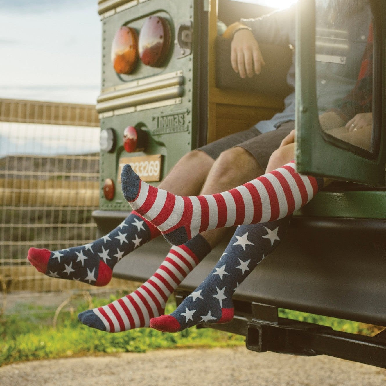 Two friends dangle their legs from the open back of a vintage green school bus at golden hour, their knee-high American Flag XL socks in VINTAGE BLUE showing red and white stripes meeting star-spangled navy panels above red toes. [lf]