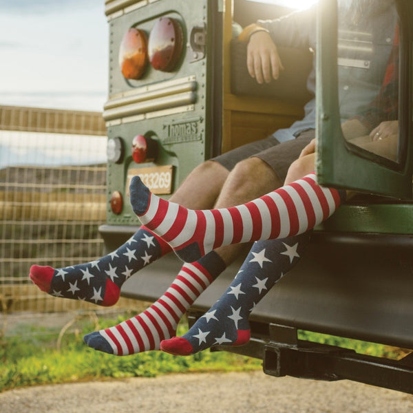 Two friends dangle their legs from the open back of a vintage green school bus at golden hour, their knee-high American Flag XL socks in VINTAGE BLUE showing red and white stripes meeting star-spangled navy panels above red toes. [lf]