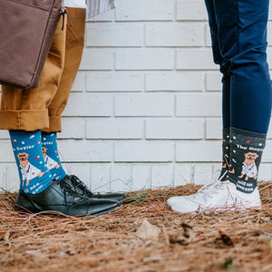 Side by side legs against a painted brick wall on autumn pine straw, paired oxford and sneaker outfits showing The Dogtor Is In socks. [lf]