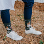 Outdoor close-up of feet in white sneakers and navy scrub pants, The Dogtor Is In socks framed by fallen leaves and pine needles. [lf]