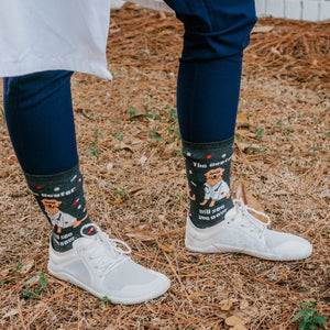 Outdoor close-up of feet in white sneakers and navy scrub pants, The Dogtor Is In socks framed by fallen leaves and pine needles. [lf]