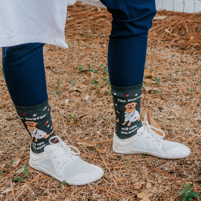 Outdoor close-up of feet in white sneakers and navy scrub pants, The Dogtor Is In socks framed by fallen leaves and pine needles. [lf]