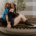 Two friends laugh back-to-back on a curved wooden city bench, hands clasped, the seated friend in black Friendship Bracelet socks striped with rainbow bands paired with black Converse high tops. [lf]
