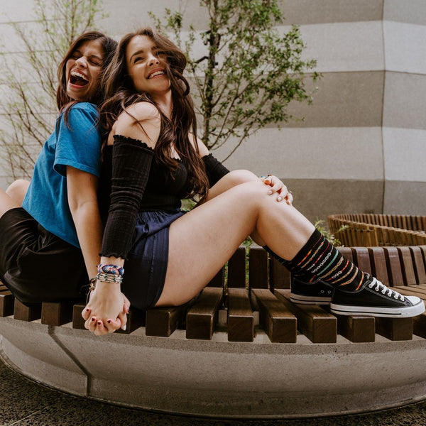 Two friends laugh back-to-back on a curved wooden city bench, hands clasped, the seated friend in black Friendship Bracelet socks striped with rainbow bands paired with black Converse high tops. [lf]