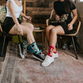 Two women relax inside a renovated vintage camper with a wood-paneled wall and a black cat painting; one wears the teal Toe Beans in Blue socks with black sneakers while a real black cat prowls the green kitchen counter behind them. [lf]