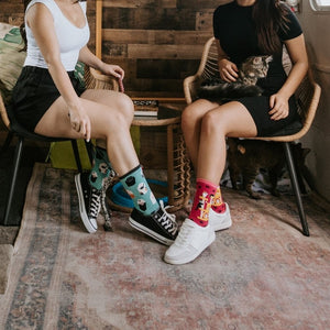 Two women relax inside a renovated vintage camper with a wood-paneled wall and a black cat painting; one wears the teal Toe Beans in Blue socks with black sneakers while a real black cat prowls the green kitchen counter behind them. [lf]