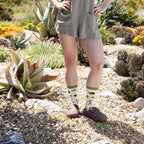 Smiling woman in olive linen romper stands hand-on-hip in a sunlit succulent garden, wearing Deserted Tortoise Bamboo socks in Ivory with brown clogs. [lf]