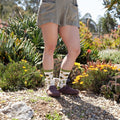 Close-up of legs walking a gravel garden path lined with yellow wildflowers and cacti, featuring Deserted Tortoise Bamboo socks in Ivory paired with brown clogs. [lf]