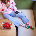 Top-down shot of a woman in a strawberry cardigan holding a takeaway coffee cup on a beige bench, a metal tray of strawberries on the table nearby, her feet crossed in pink Strawberry Threads socks in Black with vivid berry illustrations. [lf]