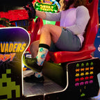 An arcade gamer grips the red blaster of a Space Invaders Frenzy cabinet, his Charge of the Robots socks in Light Gray Heather lined with chunky cartoon robots showing above black sneakers. [lf]
