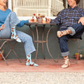 Bright outdoor seating with two coffee dates exchanging plants, the womans leg lifted to highlight Social Butterfly socks in Hemp Heather featuring orange butterflies fluttering across a soft heathered knit. [lf]