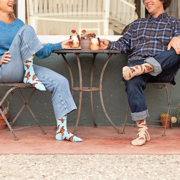 Bright outdoor seating with two coffee dates exchanging plants, the womans leg lifted to highlight Social Butterfly socks in Hemp Heather featuring orange butterflies fluttering across a soft heathered knit. [lf]