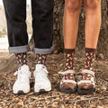 Two pairs of feet stand on a leafy forest floor beside a foraging basket of mushrooms, both wearing Colorful Caps Bamboo in Charcoal Heather socks printed with toadstools. [lf]