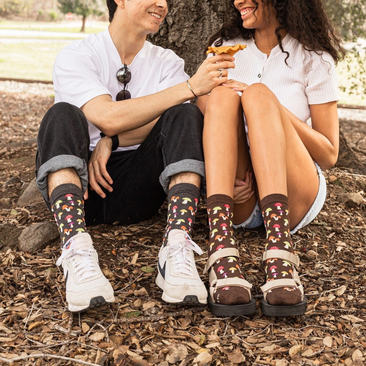 Couple laughs together at the base of an oak tree sharing a chanterelle, the pair wearing Colorful Caps Bamboo in Charcoal Heather socks with cuffed pants and shorts. [lf]
