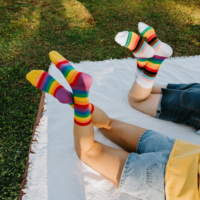 Two friends lie on a white picnic blanket in a sunny park, their crossed legs aimed skyward to show off rainbow Progress Stripes and Pride Stripes socks. [lf]