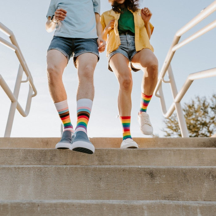 Captured mid-stride descending a staircase, a couple in shorts hops down with Progress Stripes and Pride Stripes socks bursting with color. [lf]