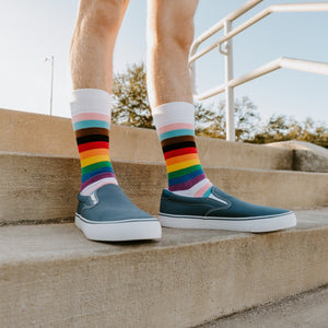 A solo close shot frames a man's feet in slate slip-ons and Progress Stripes socks, pink, white, light blue, brown, black and rainbow bands stacked above the cuff. [lf]