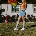 A woman in an orange tank top and denim shorts stands on a sunlit lawn holding a rainbow diamond kite, wearing Blowing In The Wind socks in Blue Heather with white sneakers in front of a pale stucco house. [lf]