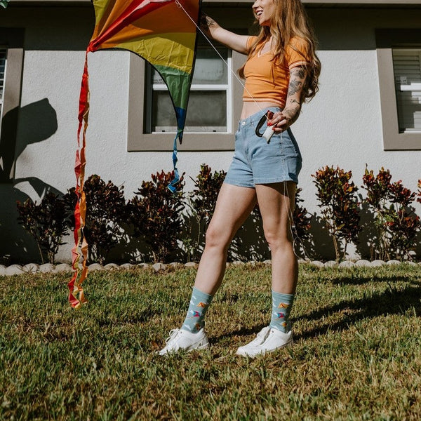 A woman in an orange tank top and denim shorts stands on a sunlit lawn holding a rainbow diamond kite, wearing Blowing In The Wind socks in Blue Heather with white sneakers in front of a pale stucco house. [lf]