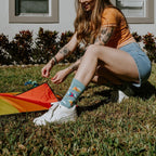 A woman kneels on a grassy yard adjusting the tail of a rainbow kite while wearing Blowing In The Wind socks in Blue Heather with white sneakers and denim shorts. [lf]