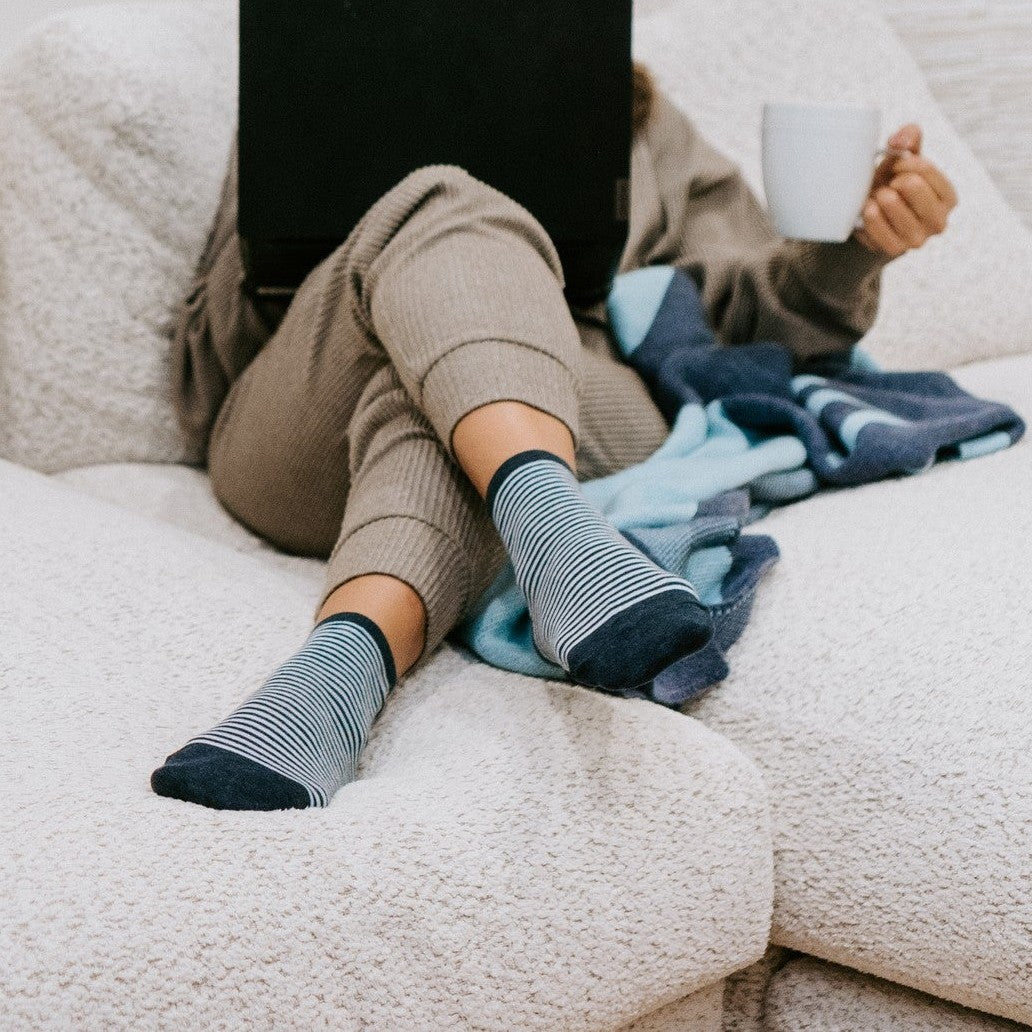 Feet propped on a cozy white couch beside a laptop and mug, Sailor Stripe socks in Navy Heather centered in the frame. [lf]