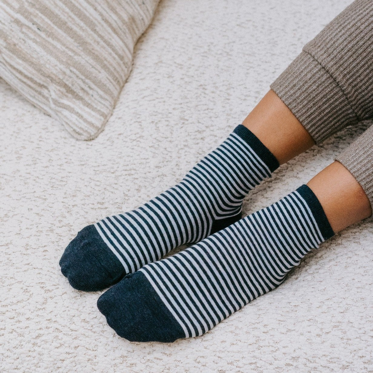 Vertical close-up of feet at a boucle cushion, Sailor Stripe socks in Navy Heather pulled neatly above the ankle. [lf]