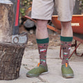 Close-up of calves in brown Are We There Yet? socks with green cuffs and tiny camper-van designs, standing on a stone patio between a chopping block and a wheelbarrow. [lf]