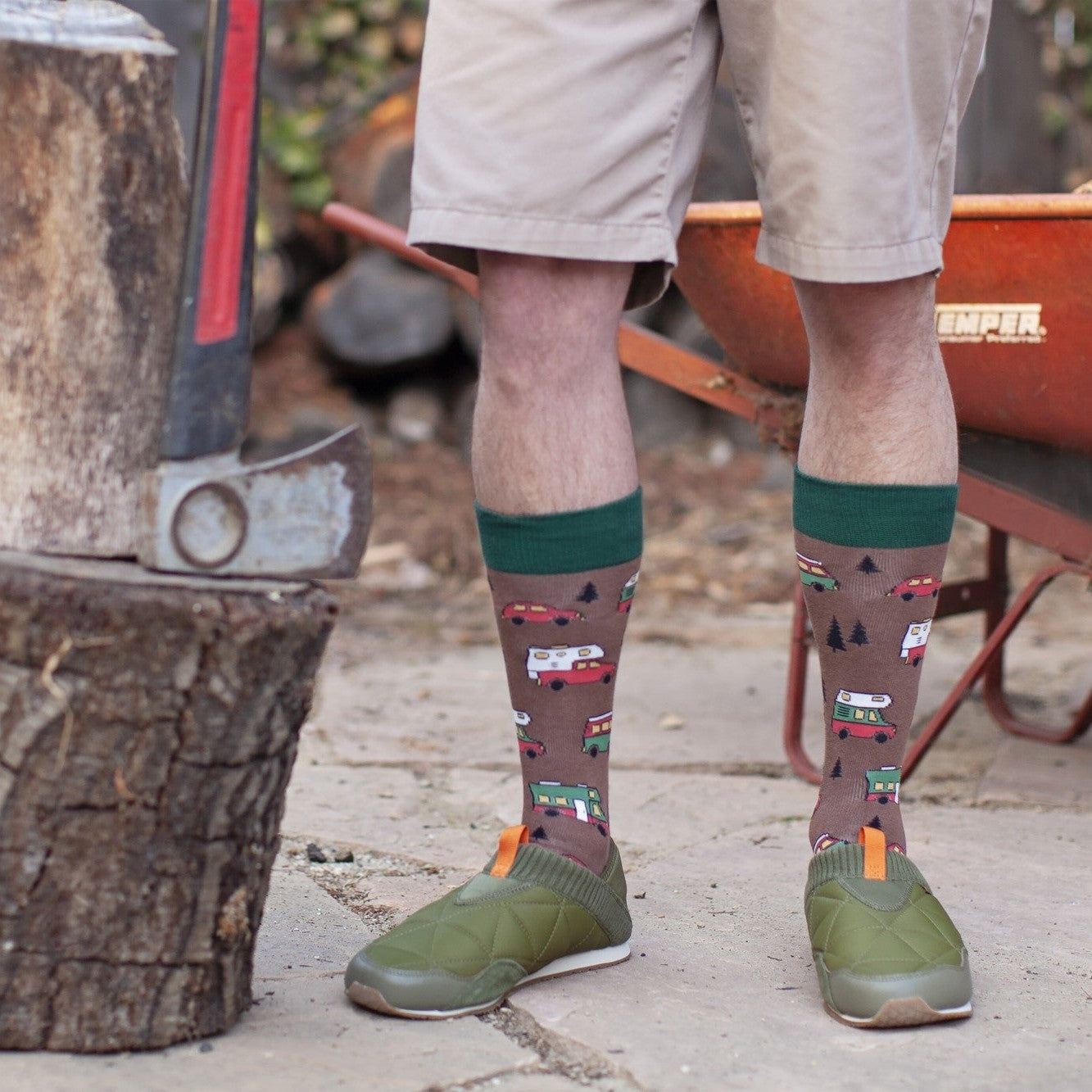 Close-up of calves in brown Are We There Yet? socks with green cuffs and tiny camper-van designs, standing on a stone patio between a chopping block and a wheelbarrow. [lf]