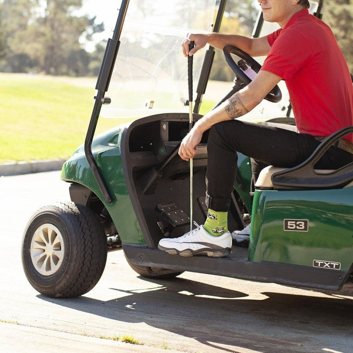 A golfer in a red polo and flat cap steers a green cart along a sun-bright fairway path, a club resting on the floor as his Putting Around socks in Green Heather flash above white sneakers. [lf]