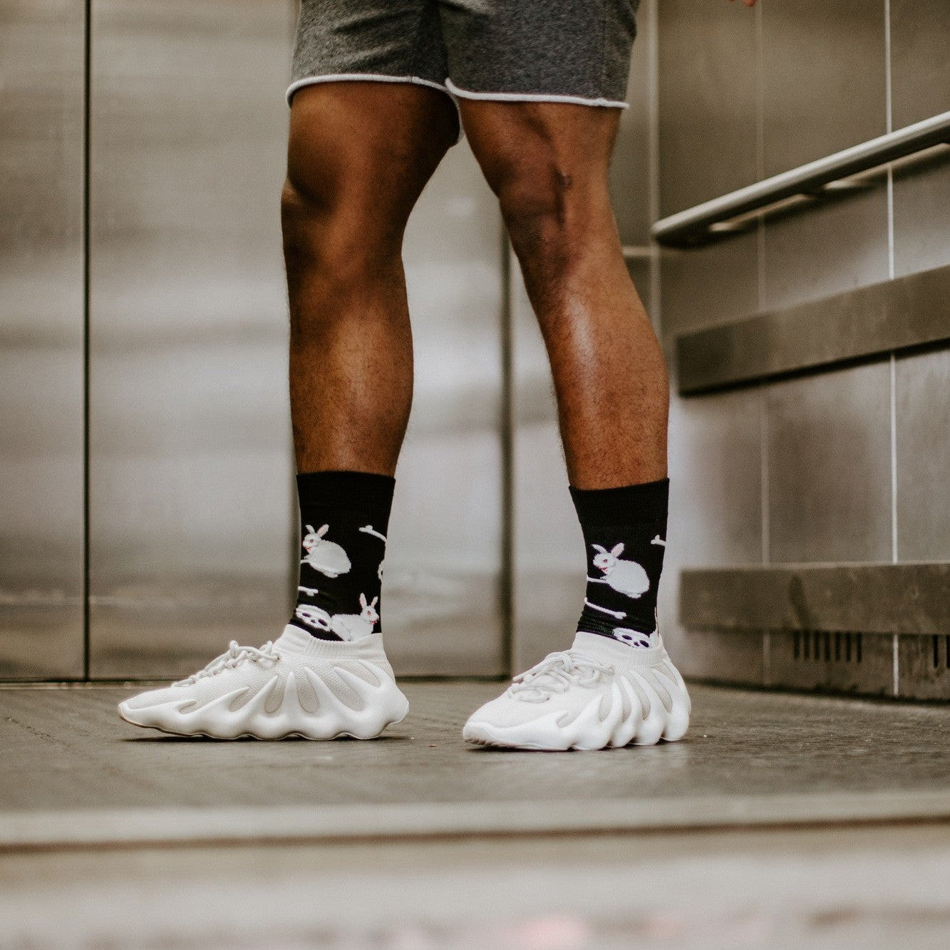A man stands inside a stainless-steel elevator in gray shorts and a light denim jacket, wearing the black Evil Bunnies socks patterned with white bunnies and crossbones above chunky white sneakers. [lf]