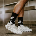 Close-up side view of the Evil Bunnies socks worn with chunky white lattice sneakers on a metal-grated floor, showing the white bunny graphics scattered across the black ribbed crew. [lf]