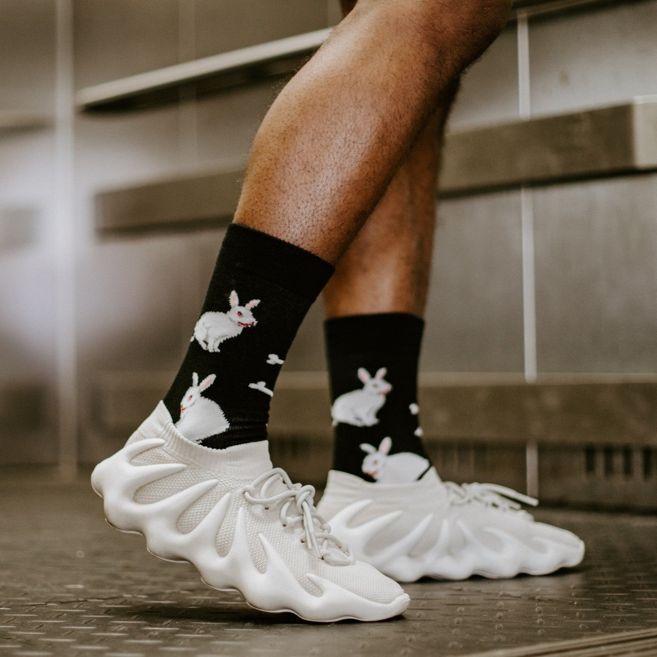 Close-up side view of the Evil Bunnies socks worn with chunky white lattice sneakers on a metal-grated floor, showing the white bunny graphics scattered across the black ribbed crew. [lf]