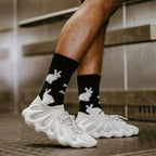 Close-up side view of the Evil Bunnies socks worn with chunky white lattice sneakers on a metal-grated floor, showing the white bunny graphics scattered across the black ribbed crew. [lf]