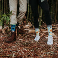 A horizontal close-up of two pairs of feet on dried bamboo leaves, with Green Thumb Bamboo in Blue socks bridging the gap between brown boots and white sneakers. [lf]
