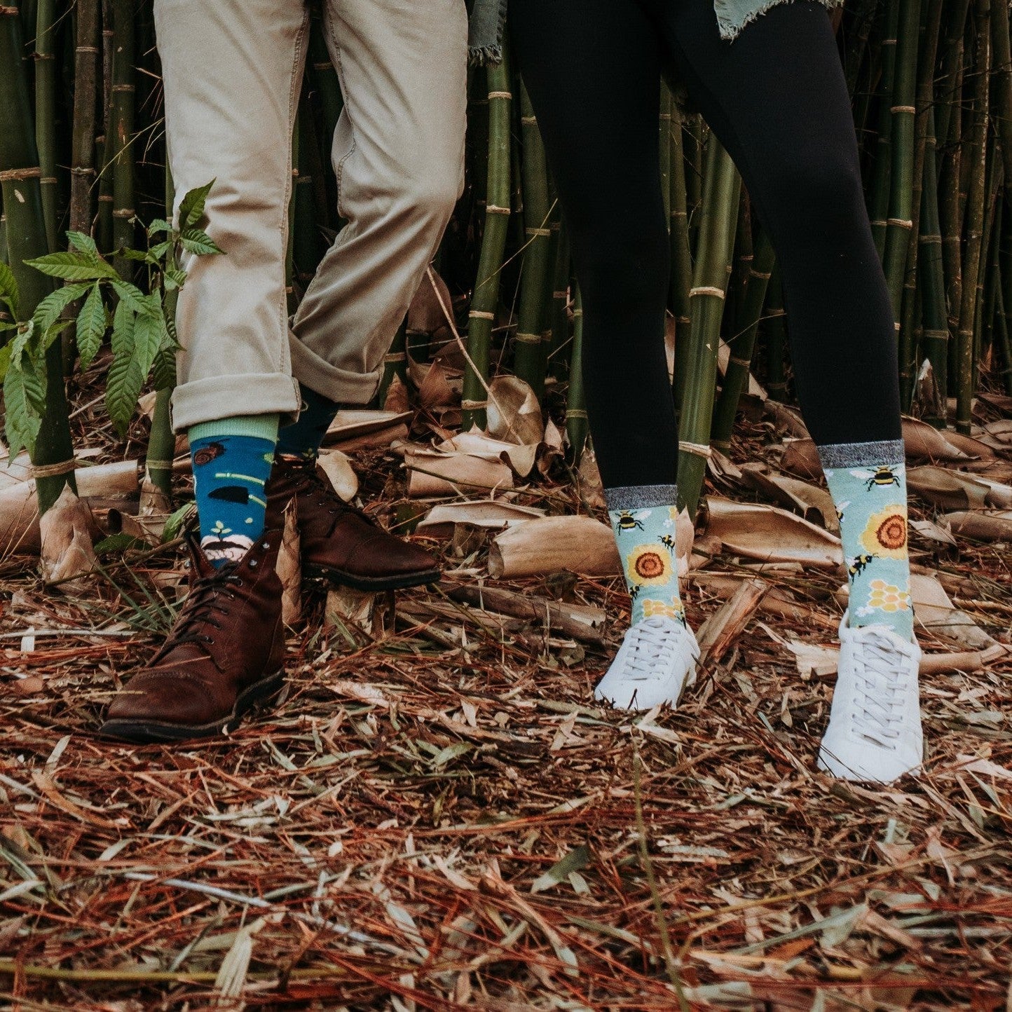 A horizontal close-up of two pairs of feet on dried bamboo leaves, with Green Thumb Bamboo in Blue socks bridging the gap between brown boots and white sneakers. [lf]