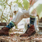 A horizontal detail of a man seated on cinder blocks in khakis and brown boots, his Green Thumb Bamboo in Blue socks featuring a rose motif against dappled light. [lf]