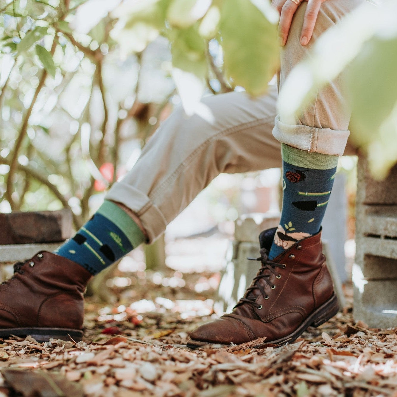 A horizontal detail of a man seated on cinder blocks in khakis and brown boots, his Green Thumb Bamboo in Blue socks featuring a rose motif against dappled light. [lf]