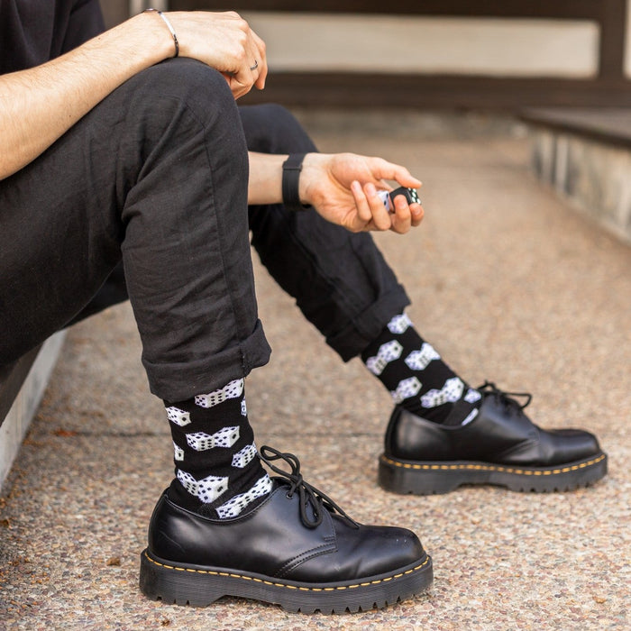 Curly-haired man in a black tee and jeans sits on a concrete step, smiling down at a die in his hand, ankles in Lucky 7's Bamboo socks in Black with allover dice graphics tucked into black platform boots. [lf]