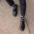 Overhead crop of a man in black jeans crossing his legs to roll a die between his hands, exposing Lucky 7's Bamboo socks in Black with bold white dice prints above chunky lace-up boots. [lf]