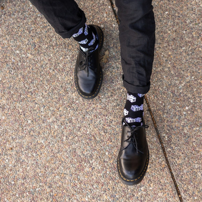 Overhead crop of a man in black jeans crossing his legs to roll a die between his hands, exposing Lucky 7's Bamboo socks in Black with bold white dice prints above chunky lace-up boots. [lf]