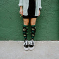 Cropped view of legs against a green stucco wall, holding a rolled cigarette, wearing knee-high Herb Garden socks in Black with black Vans sneakers. [lf]