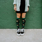 Cropped view of legs against a green stucco wall, holding a rolled cigarette, wearing knee-high Herb Garden socks in Black with black Vans sneakers. [lf]