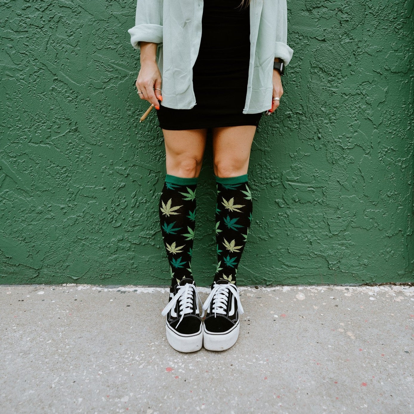 Cropped view of legs against a green stucco wall, holding a rolled cigarette, wearing knee-high Herb Garden socks in Black with black Vans sneakers. [lf]