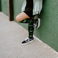 Woman leans casually against a green stucco wall in a sage shirt and black dress, wearing Herb Garden socks in Black with black Vans. [lf]