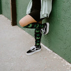 Woman leans casually against a green stucco wall in a sage shirt and black dress, wearing Herb Garden socks in Black with black Vans. [lf]