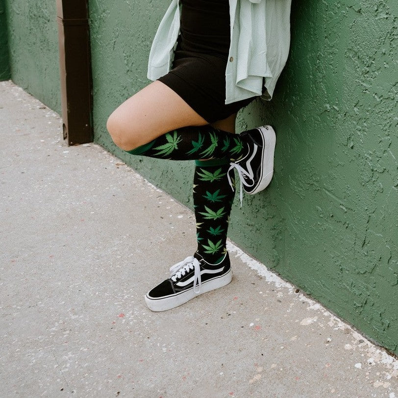Woman leans casually against a green stucco wall in a sage shirt and black dress, wearing Herb Garden socks in Black with black Vans. [lf]