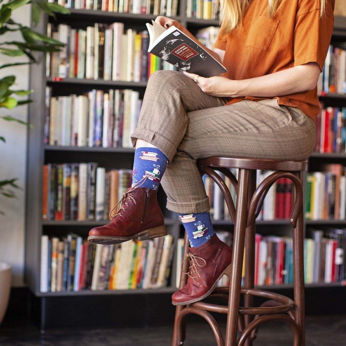 Woman in an orange tee and plaid trousers turns a page on a barstool, displaying Love Stories in Blue Heather socks with book and coffee-cup motifs above oxblood lace-up boots. [lf]