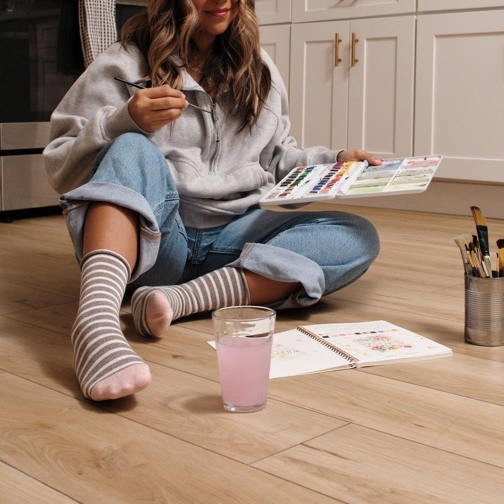 A woman sits cross-legged on a kitchen floor reviewing a watercolor palette, her Sailor Stripe Bamboo in Light Gray Heather socks paired with cuffed jeans. [lf]